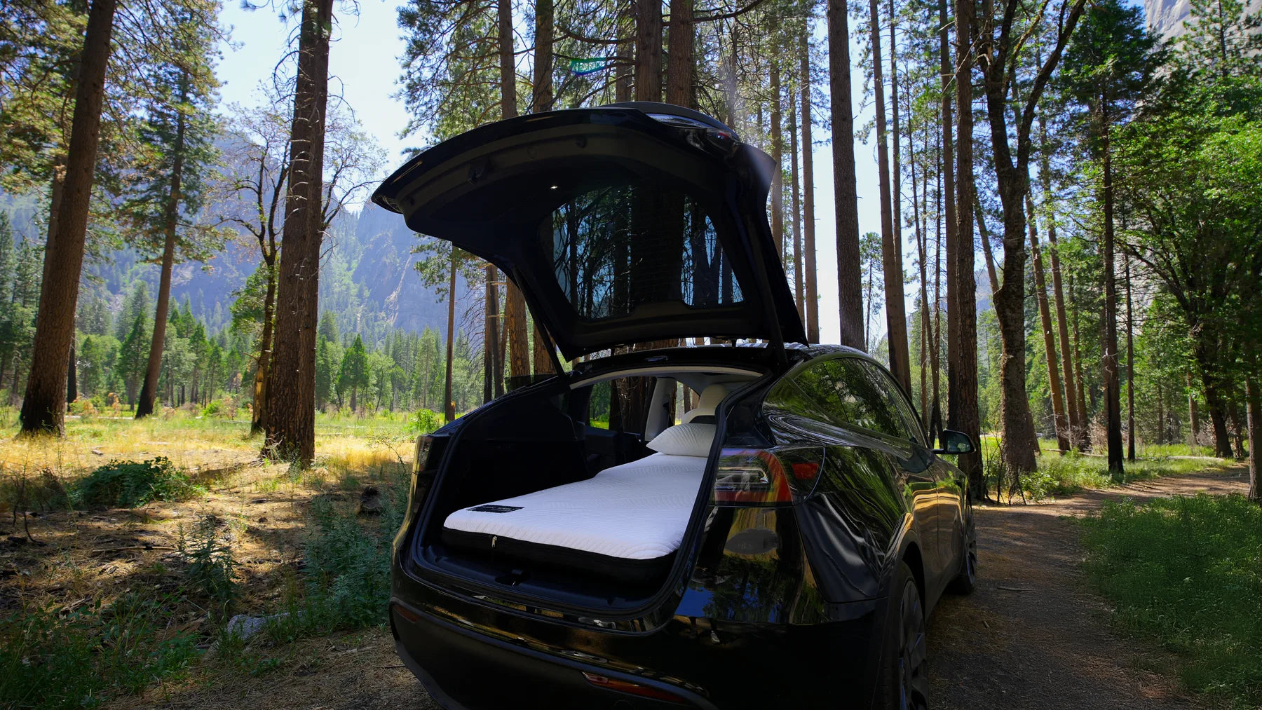 Open rear hatch of a dark Tesla with a white mattress and pillows at a forest campsite