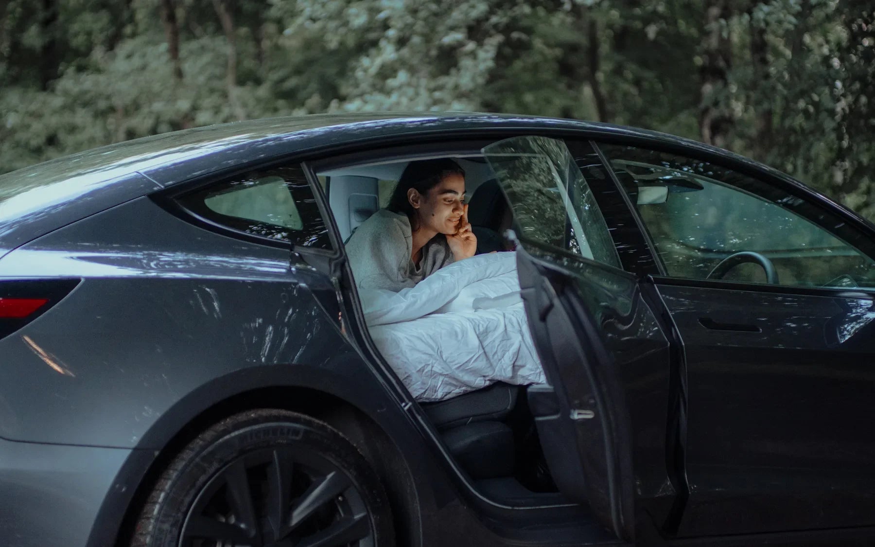 Tesla Model 3 side-door sleep setup with bedding in a wooded campsite