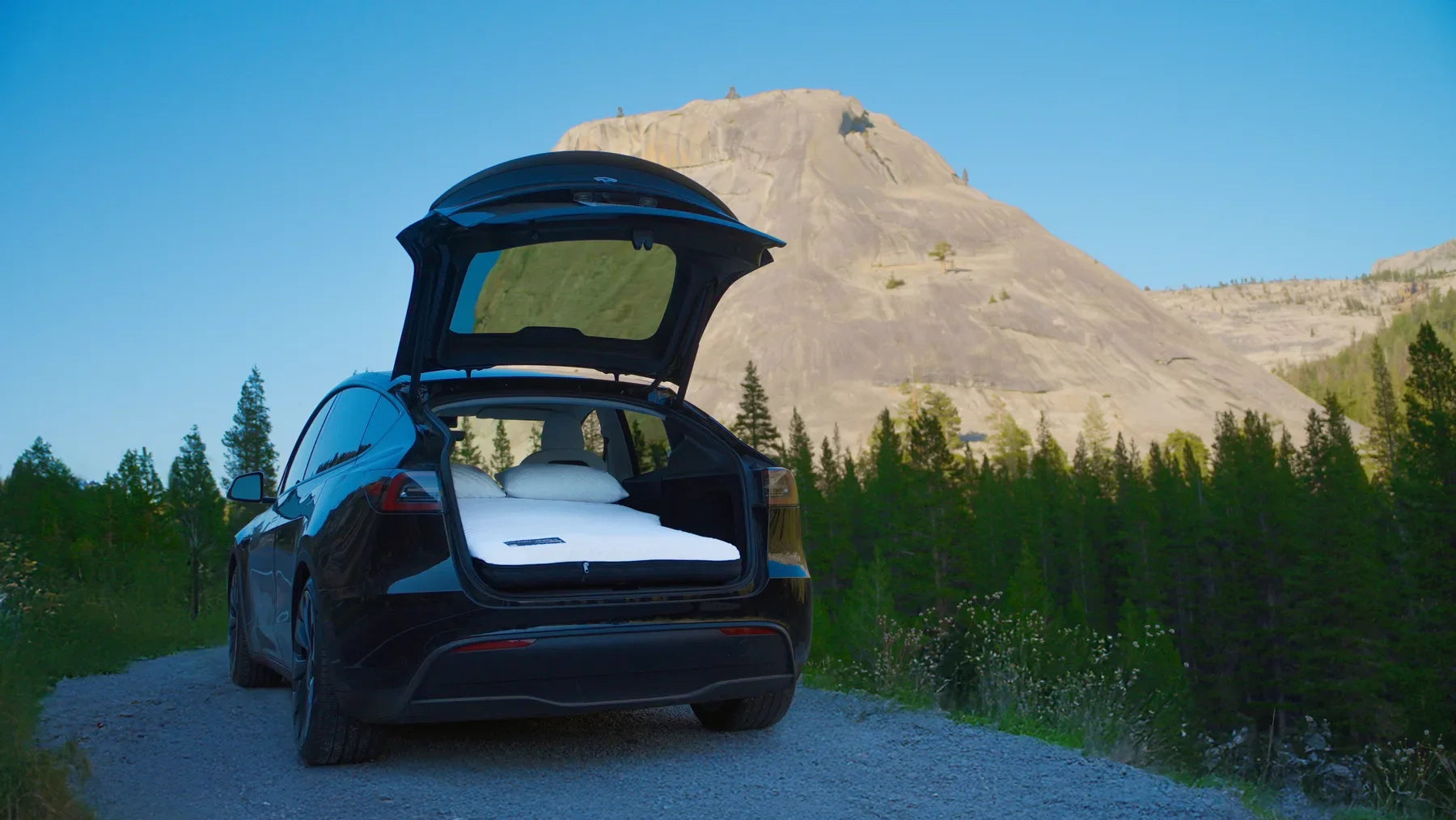 Open Tesla hatch with a two-person mattress and pillows in a mountain landscape