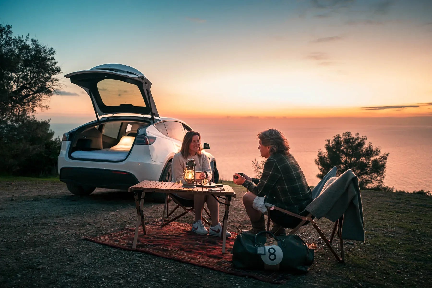Two campers beside an open Tesla with a lit rear sleep area at sunset